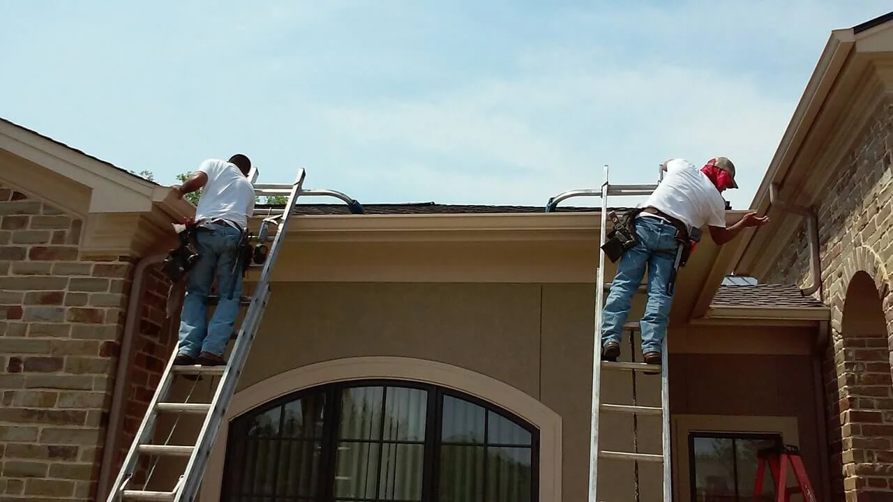 Two workers on ladders installing gutters along the roofline of a brick home