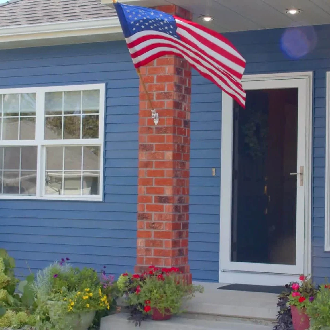 Blue lap-sided house with white grid windows, a brick porch column, an American flag and a white storm door