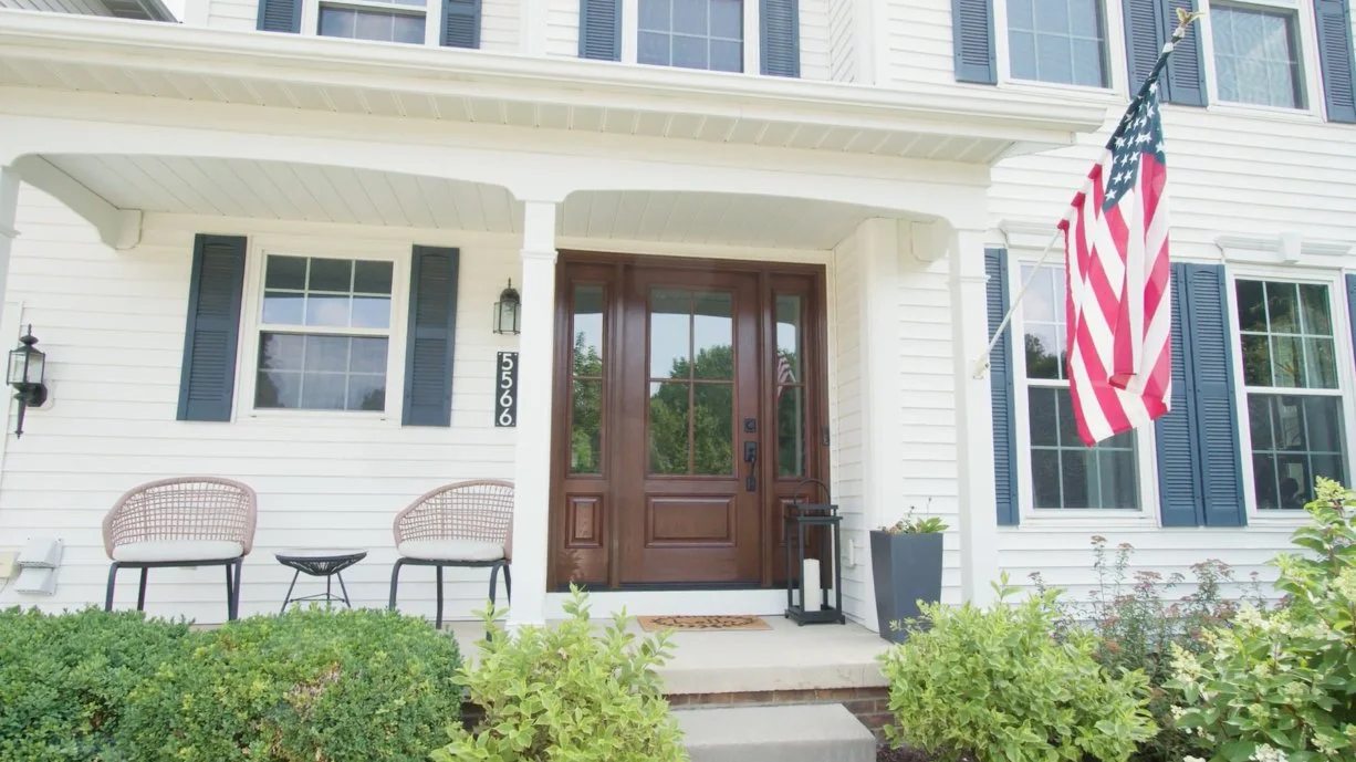 White home with a wood front door, porch chairs and an American flag