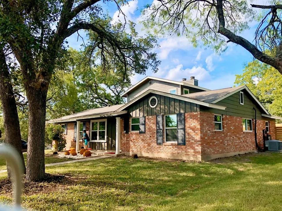 Brick ranch house with green board-and-batten siding, white windows and dark shutters under large shade trees