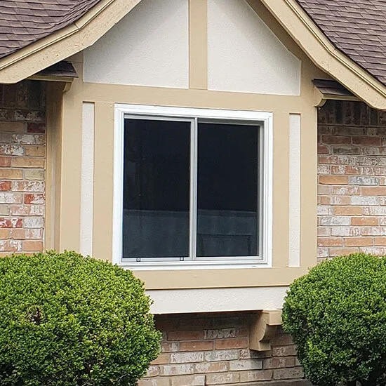 White-framed sliding window under a small gable with tan trim, set in a brick wall between two round shrubs