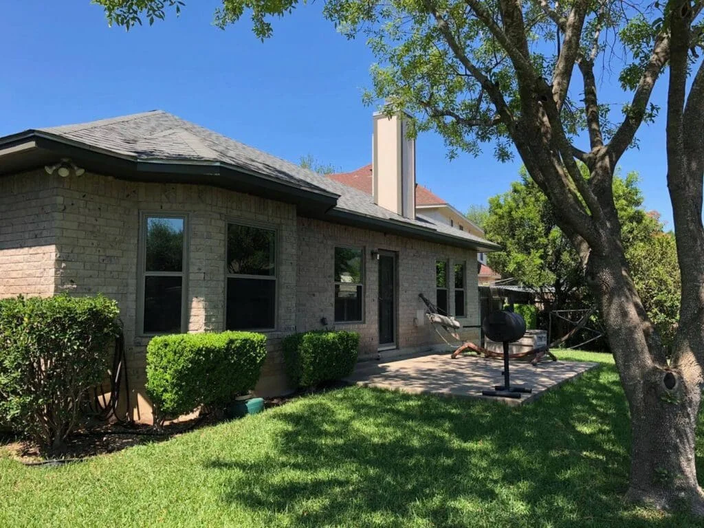 Back of a one-story tan brick house with bronze-framed windows, a patio and a large shade tree